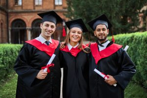 Portrait of three smiling graduate friends in graduation robes in university campus with diploma.